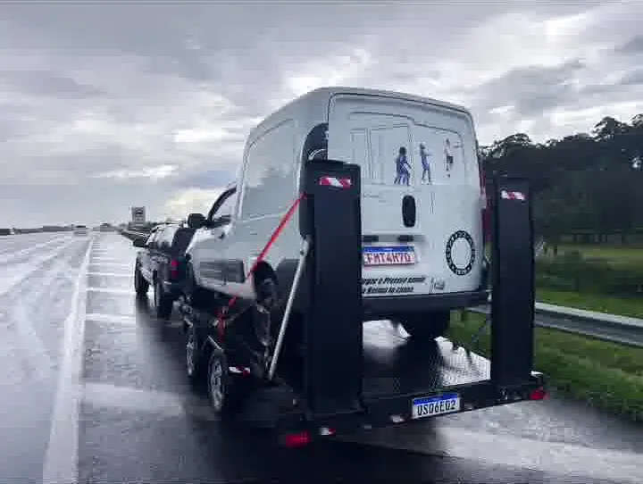 Van branca presa por cintas sobre o reboque-plataforma numa rodovia em dia de chuva